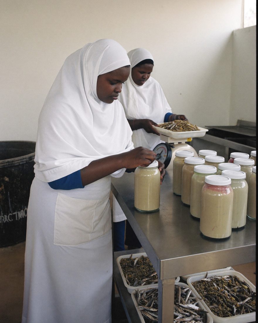 A Somali woman in an apron fills and seals jars of processed food at a stainless steel bench.