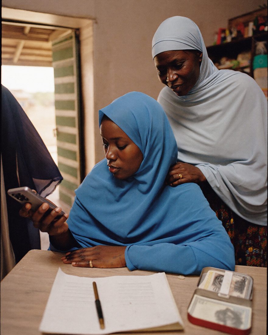 A young Somali woman completes a mobile money transaction beside a paper ledger while an older woman watches.