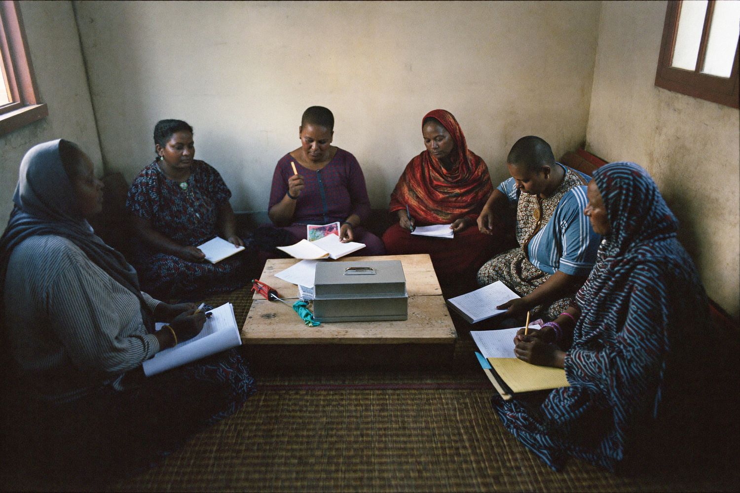 Eight Somali women sit in a circle around a savings group cash box and ledgers, recording contributions.