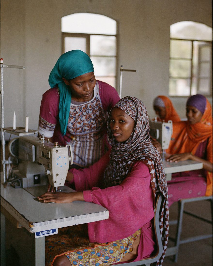 A Somali trainer leans in to guide a young woman working at an industrial sewing machine, with other trainees at machines behind them.