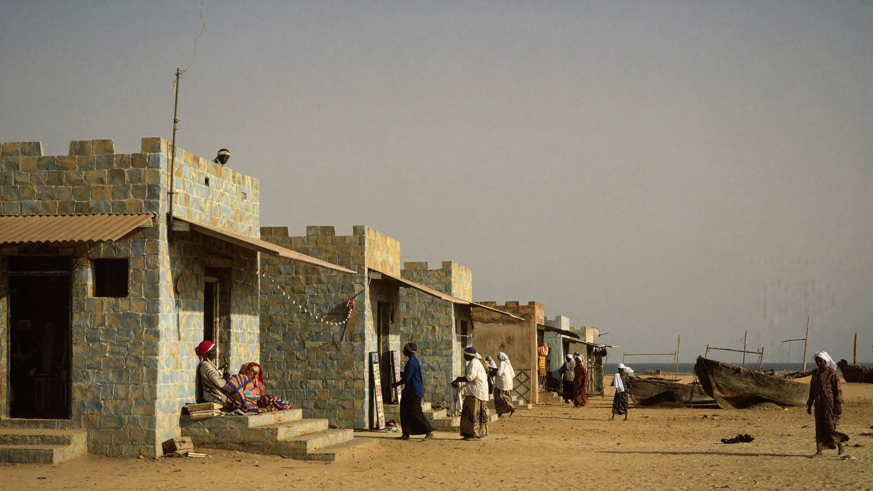 A Somali coastal town in late afternoon light, with women trading at small shopfronts along a sandy street and fishing boats beyond.