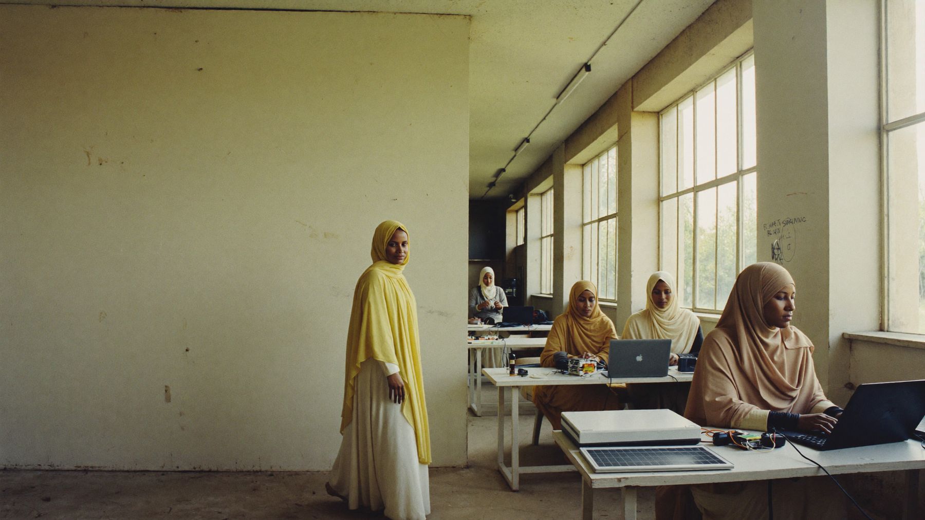 Somali women work at benches in a technical training centre, using laptops and assembling solar equipment while a trainer walks between them.