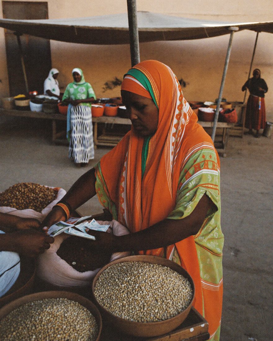 A Somali woman grain trader takes payment from a customer across baskets of sorghum and maize at a covered market.