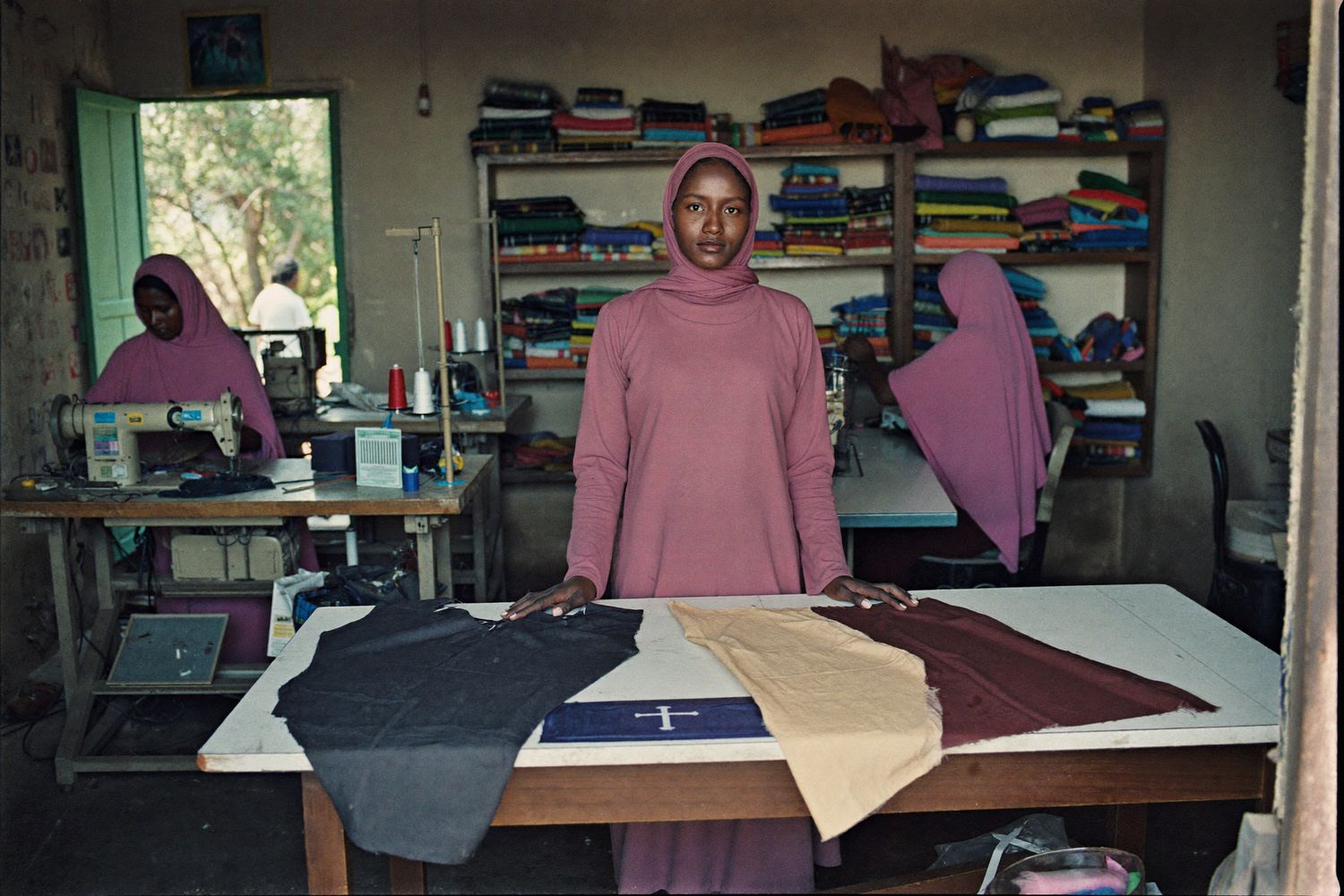A Somali woman who owns a tailoring workshop checks finished garments at a cutting table while two employees work at sewing machines behind her.