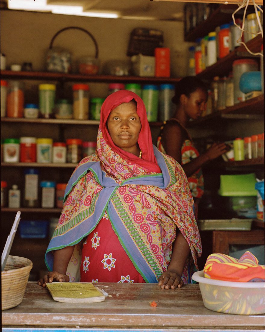A Somali woman stands behind the counter of the small shop she owns, with stocked shelves behind her.