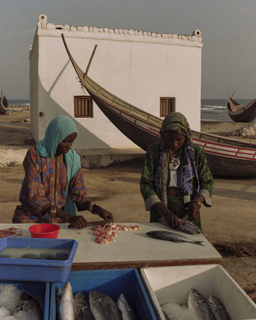 Two Somali women clean and prepare fish at an outdoor processing table beside a beached fishing boat.