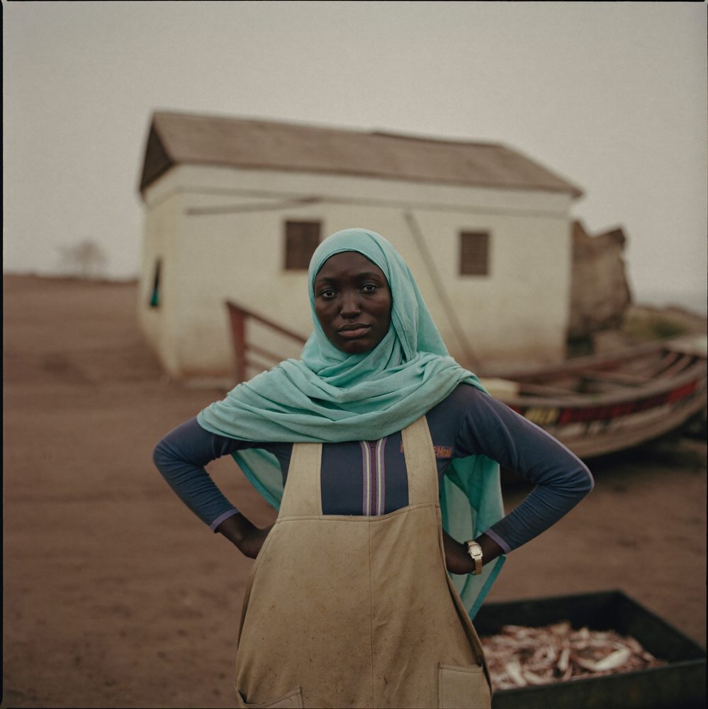 Portrait of a young Somali woman in a work apron and teal hijab standing at a coastal fish processing yard.